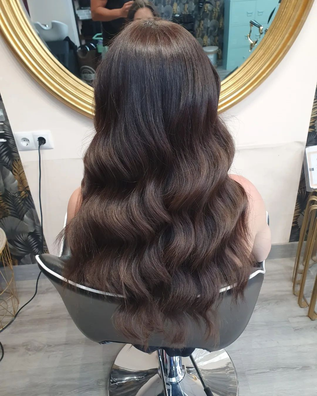 Persona sentada de espaldas en un salón de belleza, con el cabello largo y castaño oscuro, peinado en ondas suaves. Se ve un espejo circular dorado al fondo.