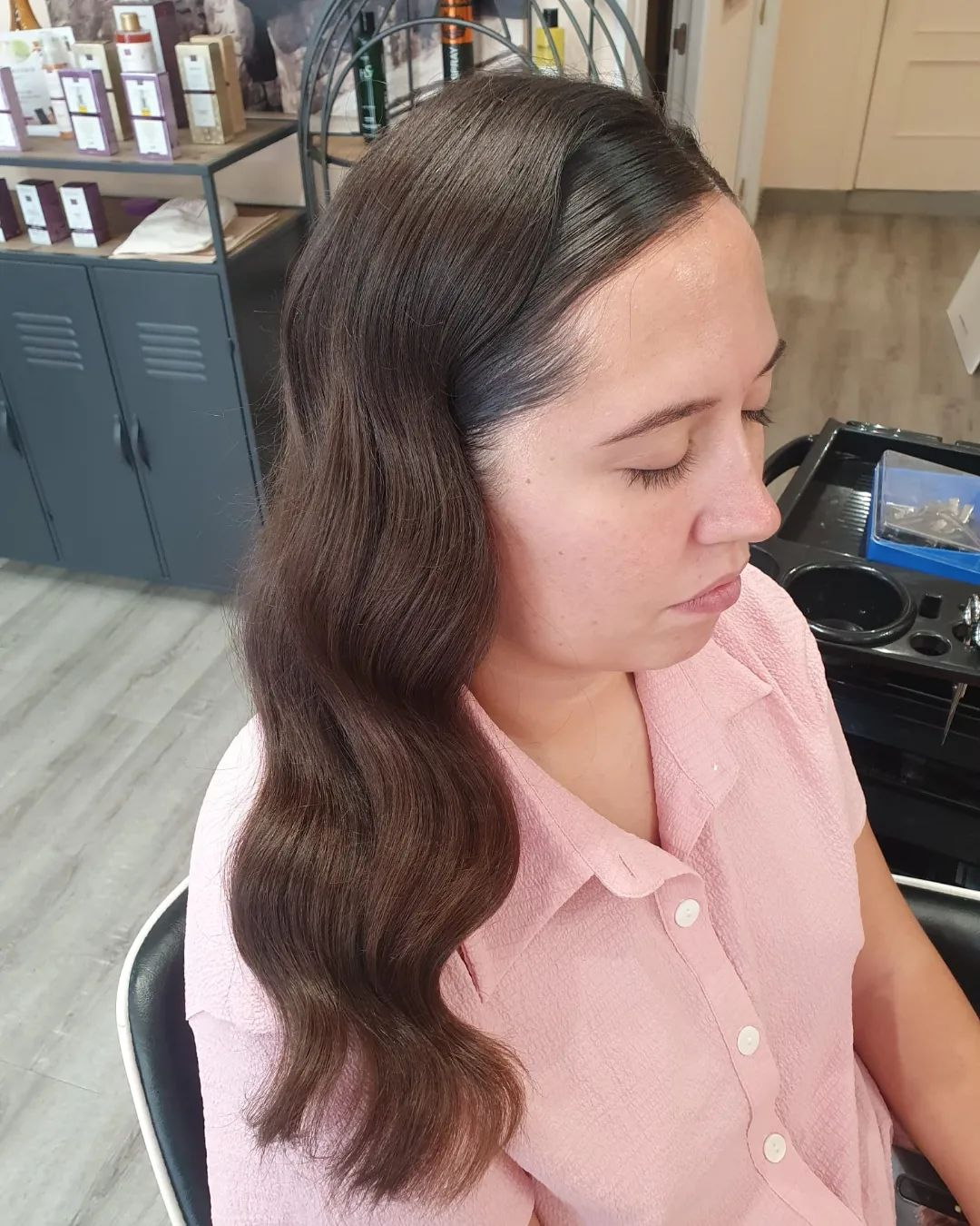 Mujer de perfil en una silla de salón, con cabello castaño oscuro, ondulado y peinado hacia un lado. Lleva una camisa rosa claro y tiene los ojos cerrados.