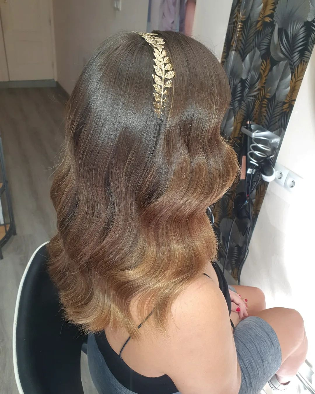 Mujer con cabello castaño largo y ondulado, adornado con una diadema dorada con diseño de hojas. Está sentada en una silla del salón.