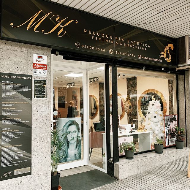 Fachada de una peluquería con letrero que dice "MK Peluquería Estética Femenina & Masculina", mostrando el interior con espejos dorados y decoración navideña.