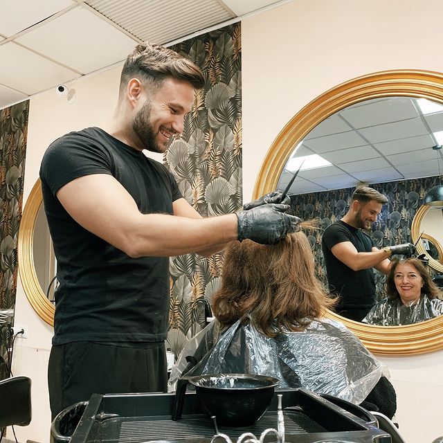 Estilista aplicando tinte en el cabello de una clienta, ambos sonrientes. El estilista lleva guantes y la clienta está cubierta con una capa plástica.