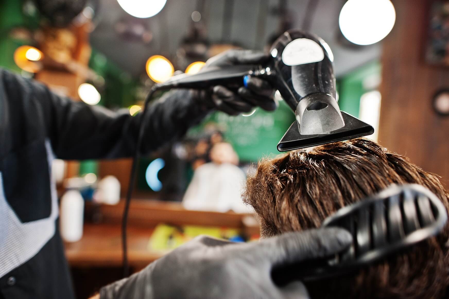 Mano de un barbero usando un secador y un cepillo mientras seca el cabello de un cliente en una barbería, con luces redondas de fondo.