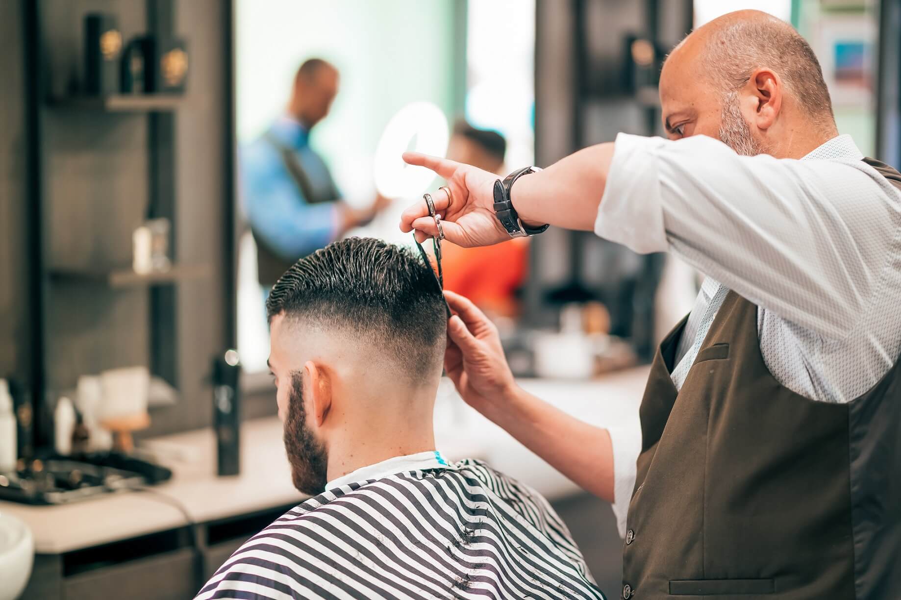 Barbero cortando el cabello de un cliente desde la parte posterior. El cliente tiene un corte estilo fade, y el barbero sostiene unas tijeras mientras trabaja.