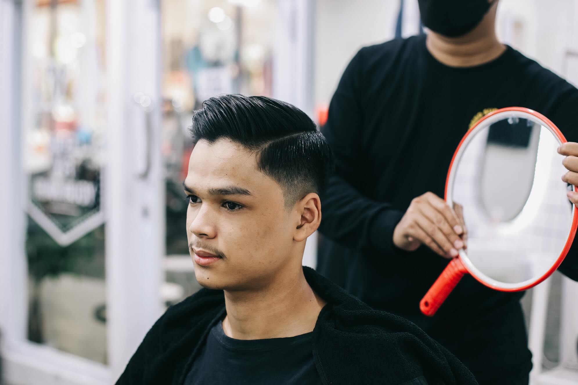 Cliente sentado en una silla de peluquería, con un peinado corto, liso y con raya lateral definida. El peluquero, de pie detrás, sostiene un espejo de mano redondo.