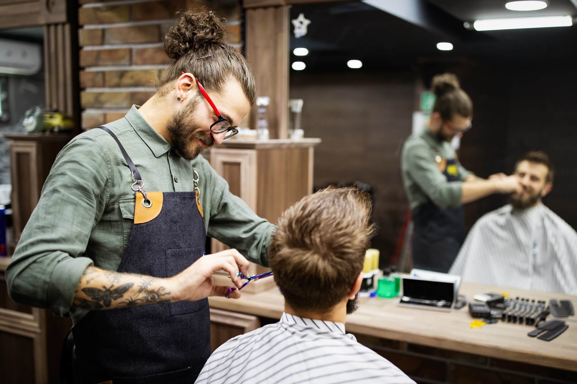 Barbero con gafas y tatuajes cortando el cabello de un cliente en una barbería. Ambos están de perfil, y se observa un espejo en la pared frente al cliente.