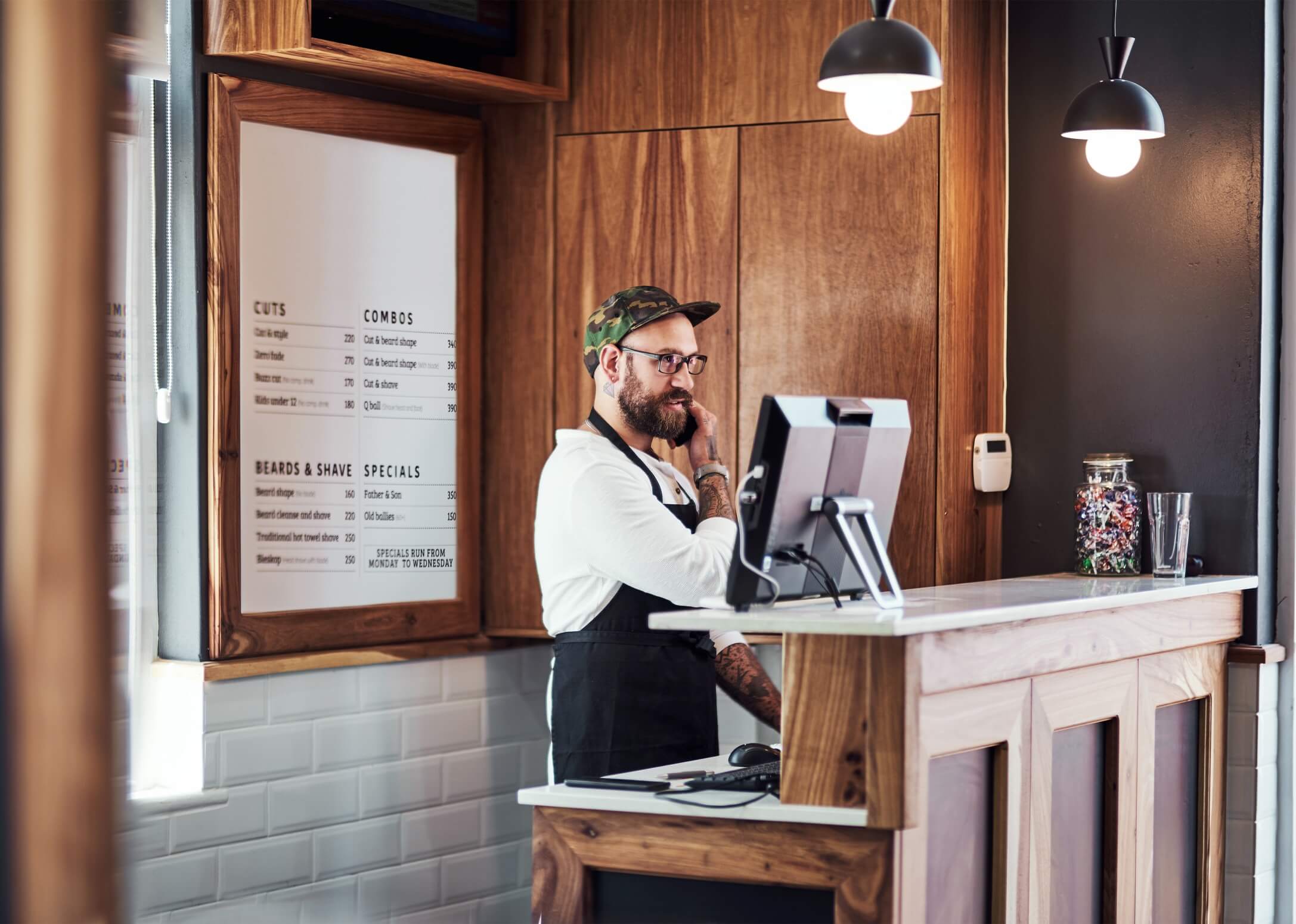 Hombre con barba y gafas atendiendo la recepción de una barbería. Está frente a una computadora, y en la pared se muestran listas de precios para cortes y servicios de barba.