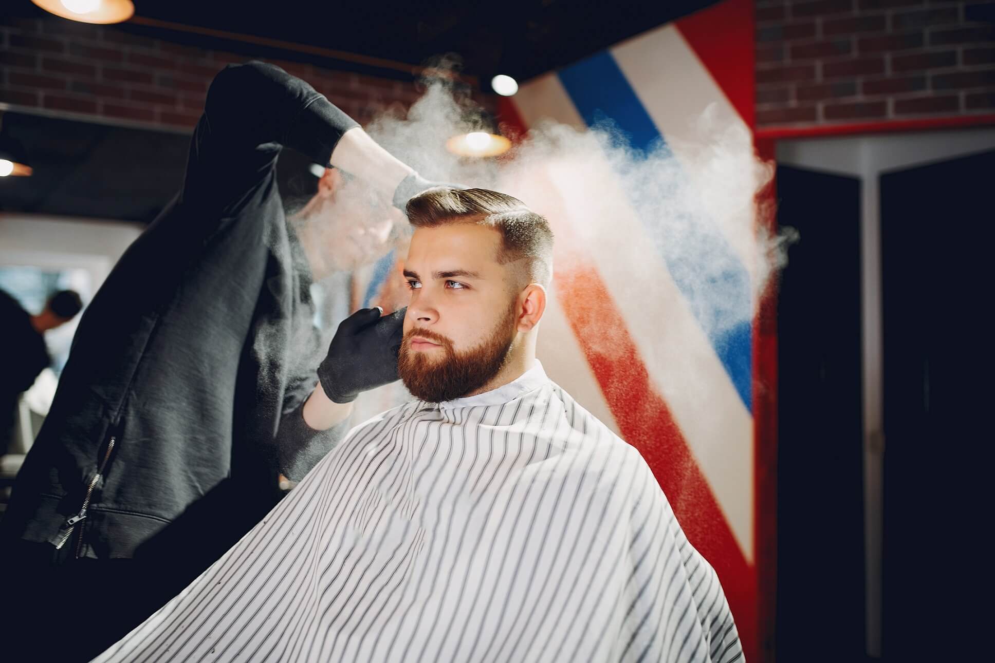 Hombre con barba y cabello corto sentado en una silla de barbería mientras un barbero aplica spray en su cabello. Al fondo se ve un poste de barbería con franjas rojas, blancas y azules.